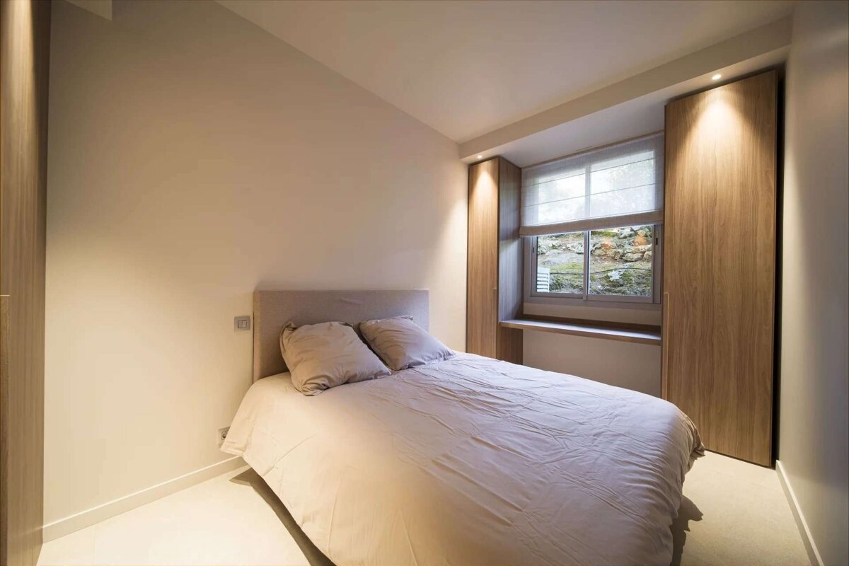 A modern, minimalist bedroom featuring a bed with light bedding, built-in wooden wardrobes flanking a window with a view of rocky greenery, and soft lighting.