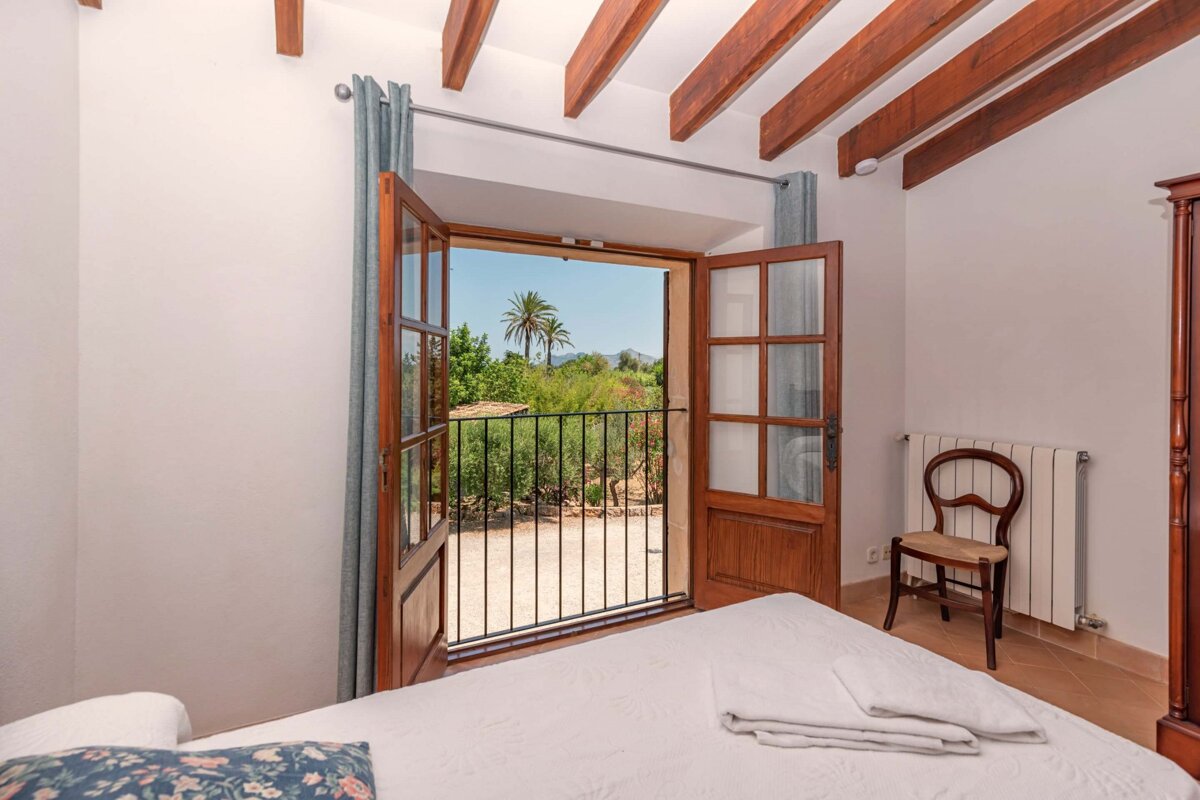 A cozy bedroom with open wooden balcony doors framing a scenic view of palm trees, mountains, and greenery. Features exposed wooden ceiling beams.