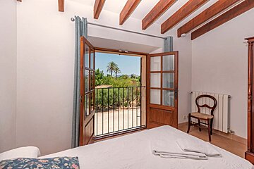 A cozy bedroom with open wooden balcony doors framing a scenic view of palm trees, mountains, and greenery. Features exposed wooden ceiling beams.