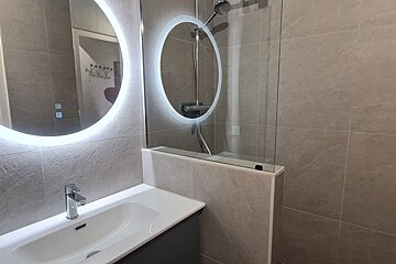 A modern grey-tiled bathroom featuring a circular LED-backlit mirror, a white sink, and a glass-paneled shower.