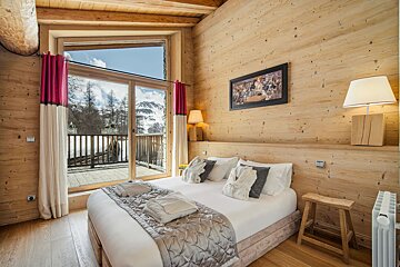 Rustic wooden chalet bedroom with a large bed, warm lighting, and glass doors opening to a scenic snowy mountain view.