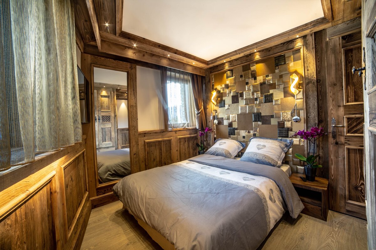 Rustic wood-paneled bedroom with a cozy bed, warm glowing lights, and a decorative shimmering gold-tiled accent wall.