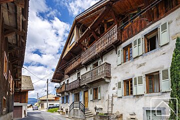 Rustic Alpine village street scene with old stone and timber buildings, wooden balconies, light shutters, and a bright blue sky with clouds.