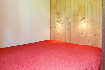 A simple bedroom with a red bedspread against a wooden paneled wall featuring two sconce lights, and an adjacent plain white wall.