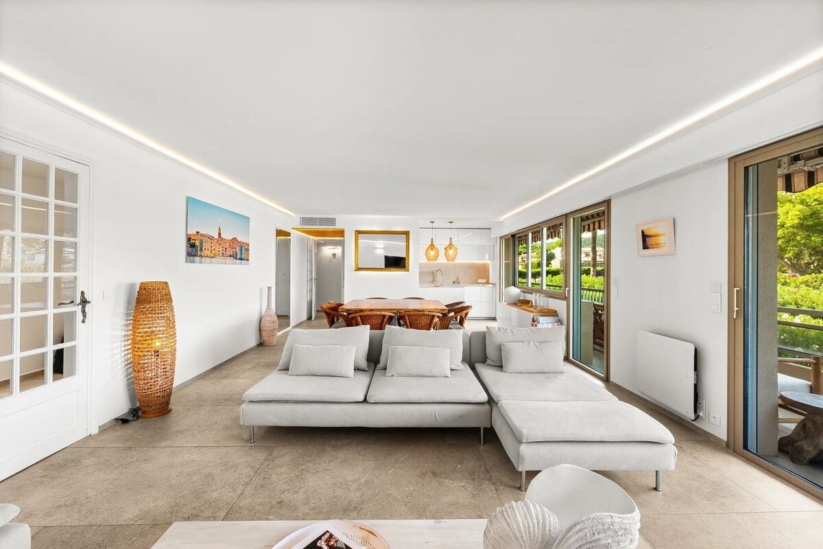 Modern, white-walled living room with a large grey sectional, tiled floor, dining area in back, and sliding doors opening to a balcony.