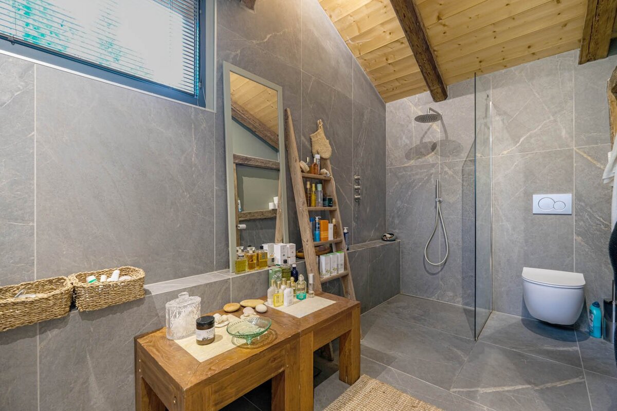 A contemporary grey tiled bathroom with a sloped wooden beam ceiling, glass shower, wall-mounted toilet, and rustic wooden vanity furniture.
