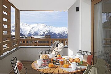A sunny balcony with breakfast (juice, croissants, bread) laid out on a table, offering a stunning panoramic view of snow-covered mountains.