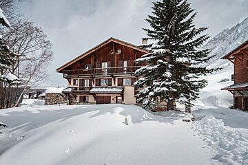 A large wooden chalet nestled in deep snow next to a snow-covered pine tree in a mountain landscape.