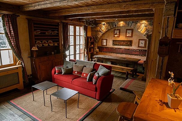 A cozy, rustic room features a red sofa, wooden beams, a stone archway, and plaid accents, creating a warm, inviting atmosphere.
