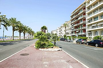 A sunny, wide street lined with palm trees and multi-story buildings, with parked cars and a clear blue sky, likely a coastal city.