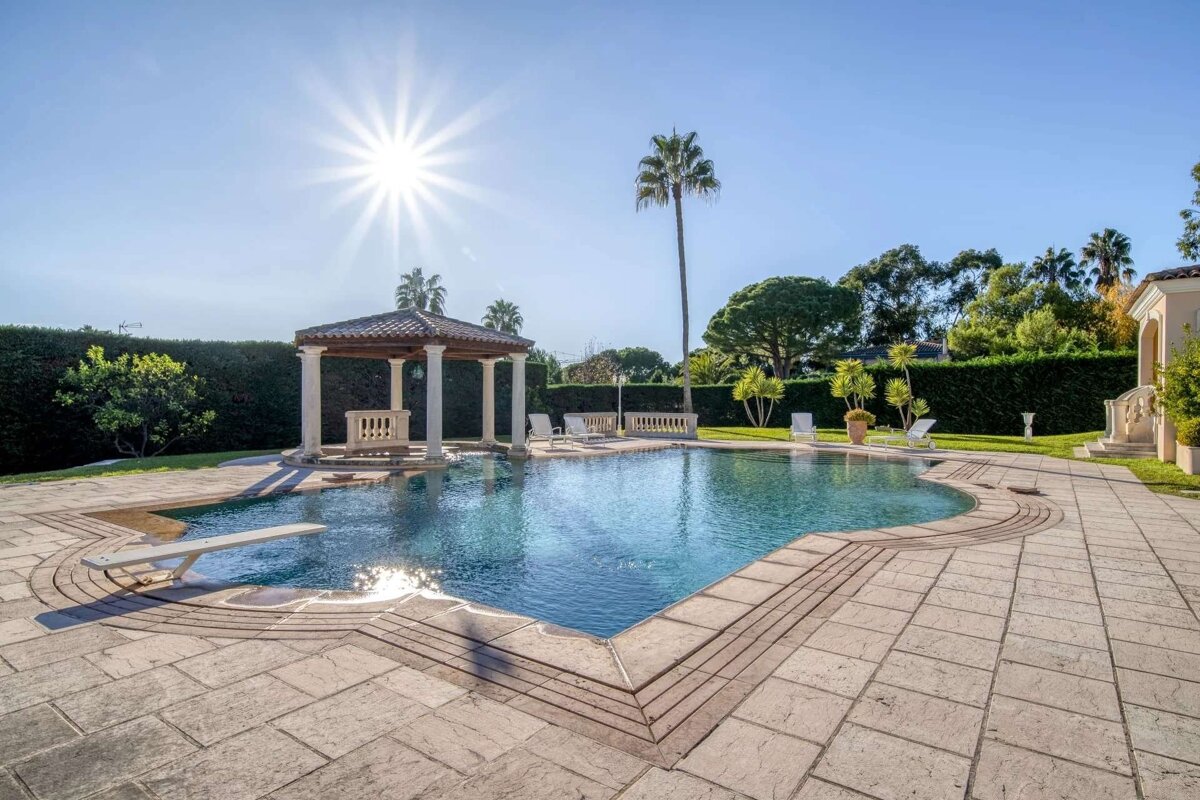 A sun-drenched, luxurious outdoor pool area with a classic gazebo, diving board, towering palm trees, and an expansive paved patio.