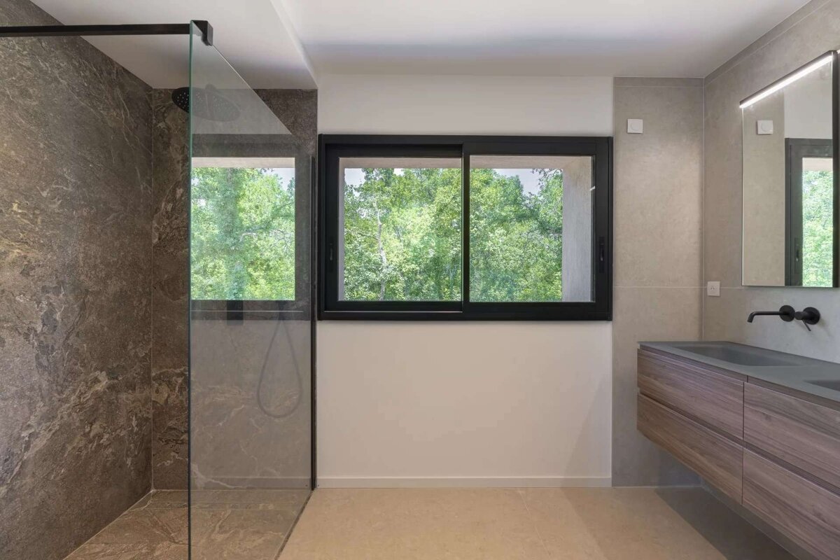 A modern bathroom features a glass shower with dark stone walls, a large window with green tree views, and a floating wooden vanity with dual sinks.