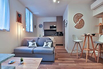 A modern open-plan studio featuring a grey sofa, compact kitchen, and a wood dining table with bar stools on a warm hardwood floor.