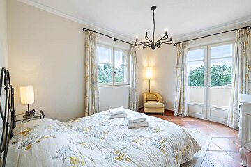 A bright bedroom with a floral quilt bed, terracotta floors, two large windows, a black chandelier, and a yellow accent chair.