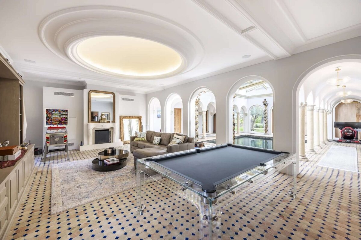 Luxurious game room with a clear pool table, patterned tile floor, fireplace, pinball machine, and arches leading to an indoor pool.