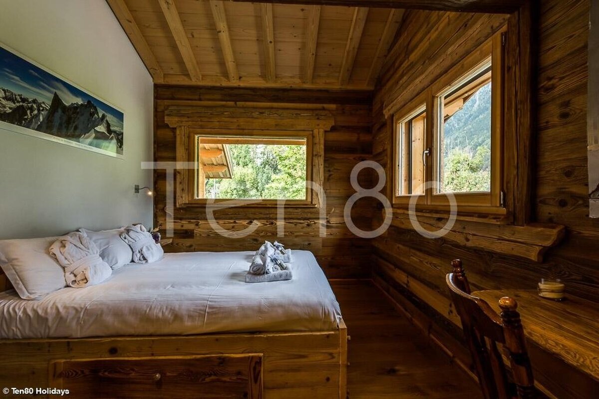 Cozy rustic bedroom in a wooden chalet with a white bed, log walls, and windows looking out onto nature.