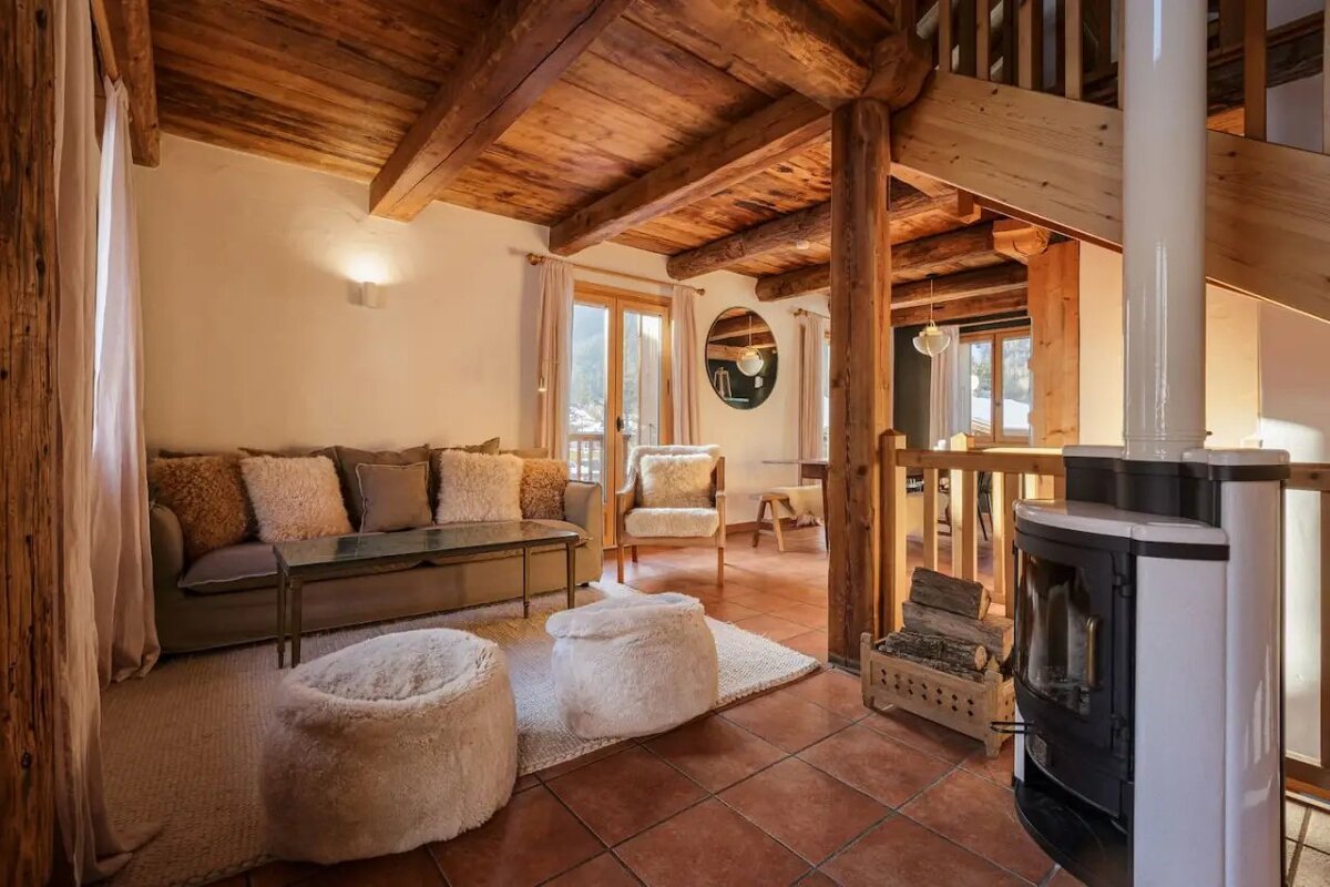 Cozy rustic cabin living room featuring exposed wooden beams, a plush sofa, fuzzy floor poufs, tiled flooring, and a modern wood stove.