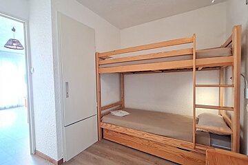 A bright room with wooden bunk beds, featuring a pull-out trundle bed, white textured walls, light wood flooring, and a closet.