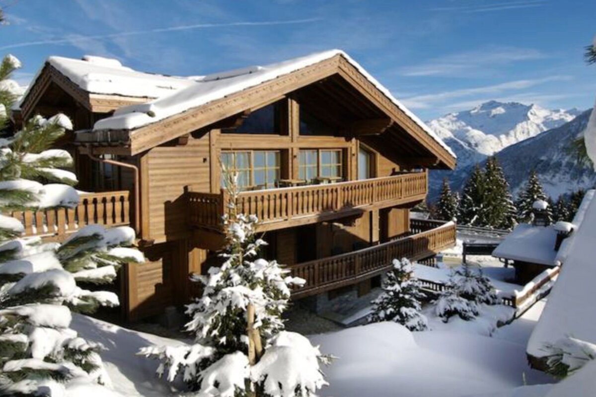 A snowy wooden chalet with balconies, surrounded by snow-laden pines, set against majestic snow-capped mountains under a clear blue sky.