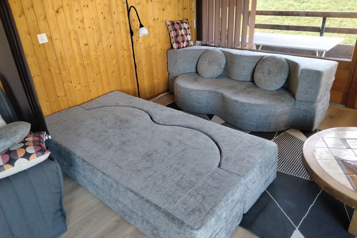 A modern living room features gray, curvy modular sofas, wood-paneled walls, and a window overlooking a balcony.