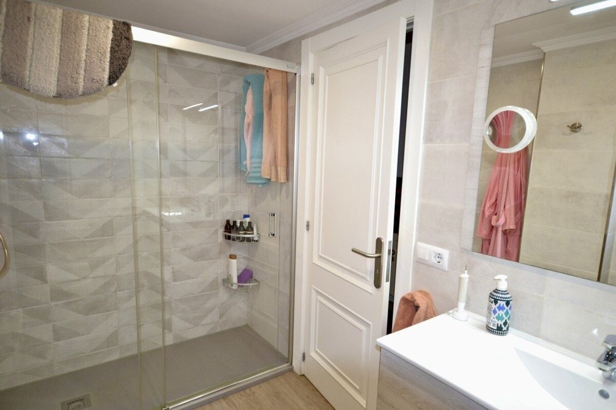 A modern bathroom features a glass shower, white door, sink vanity, and a large mirror reflecting a pink bathrobe and towels.