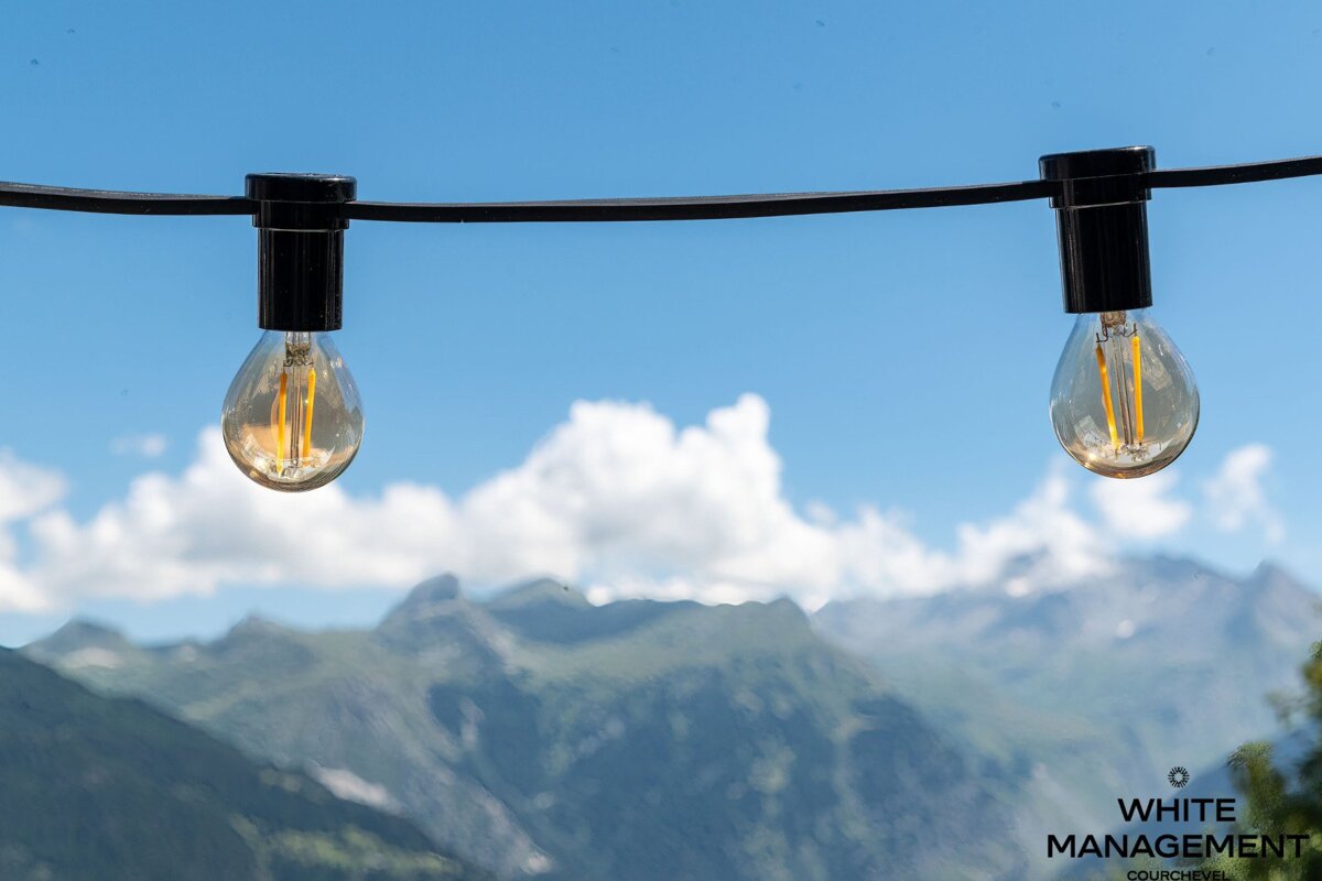 Two string lights hang against a vibrant blue sky with white clouds, overlooking blurred green mountains.