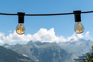 Two string light bulbs hang against a blurred backdrop of mountains and a bright blue sky.