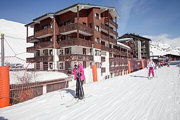 Two skiers descend a snowy slope past large chalet apartments and safety nets. A group of children ski behind. Bright, sunny mountain resort scene.
