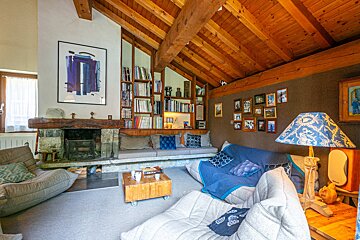 An inviting chalet-style living room with a stone fireplace, beamed ceiling, built-in bookshelves, comfortable seating, and framed pictures.