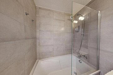 Modern bathroom featuring a white bathtub with a glass screen, chrome shower head, and large beige tiled walls.