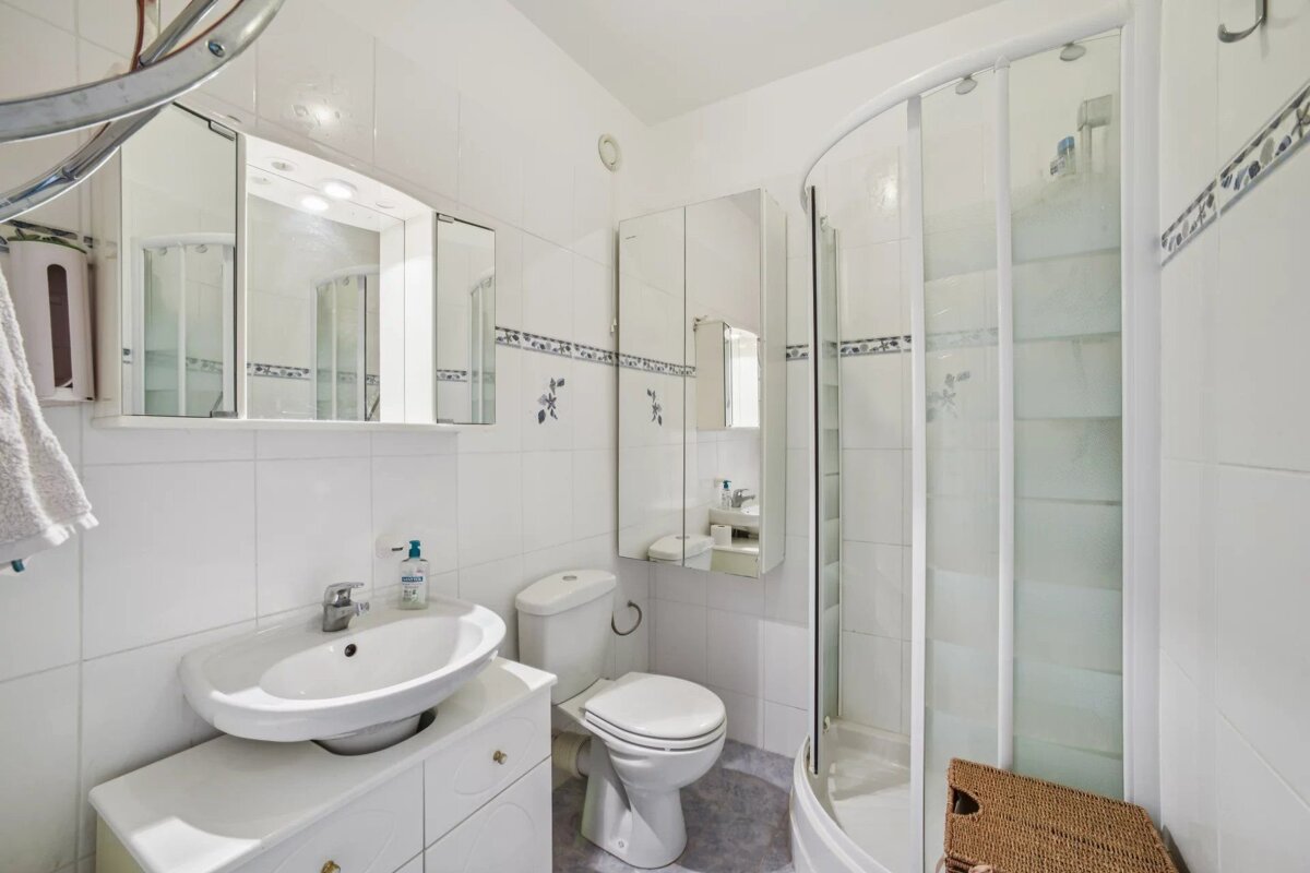 A bright, white tiled bathroom featuring a sink, toilet, corner shower, multiple mirrors, and blue decorative tile accents.