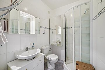A bright, white tiled bathroom featuring a sink, toilet, corner shower, multiple mirrors, and blue decorative tile accents.