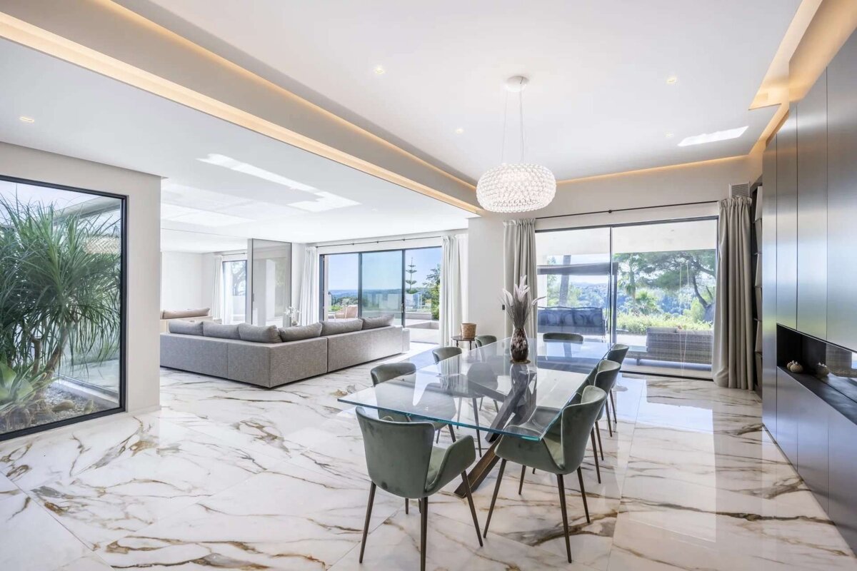 Elegant open-plan living/dining room with stunning marble floors, glass table, gray sofa, and panoramic windows offering scenic outdoor views.