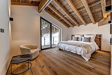 Cozy bedroom with exposed wooden beams, hardwood floor, a large window overlooking a snowy landscape, and a comfortable bed.