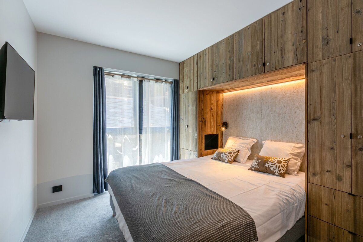 A cozy bedroom features rustic wood paneling, a bed with snowflake pillows, a window with sheer curtains, and a wall-mounted TV. Warm backlighting enhances the ambiance.