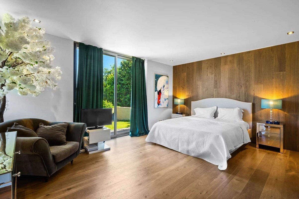 Bright, modern bedroom with a white bed, wood paneling, large windows overlooking a garden, and a cherry blossom tree.