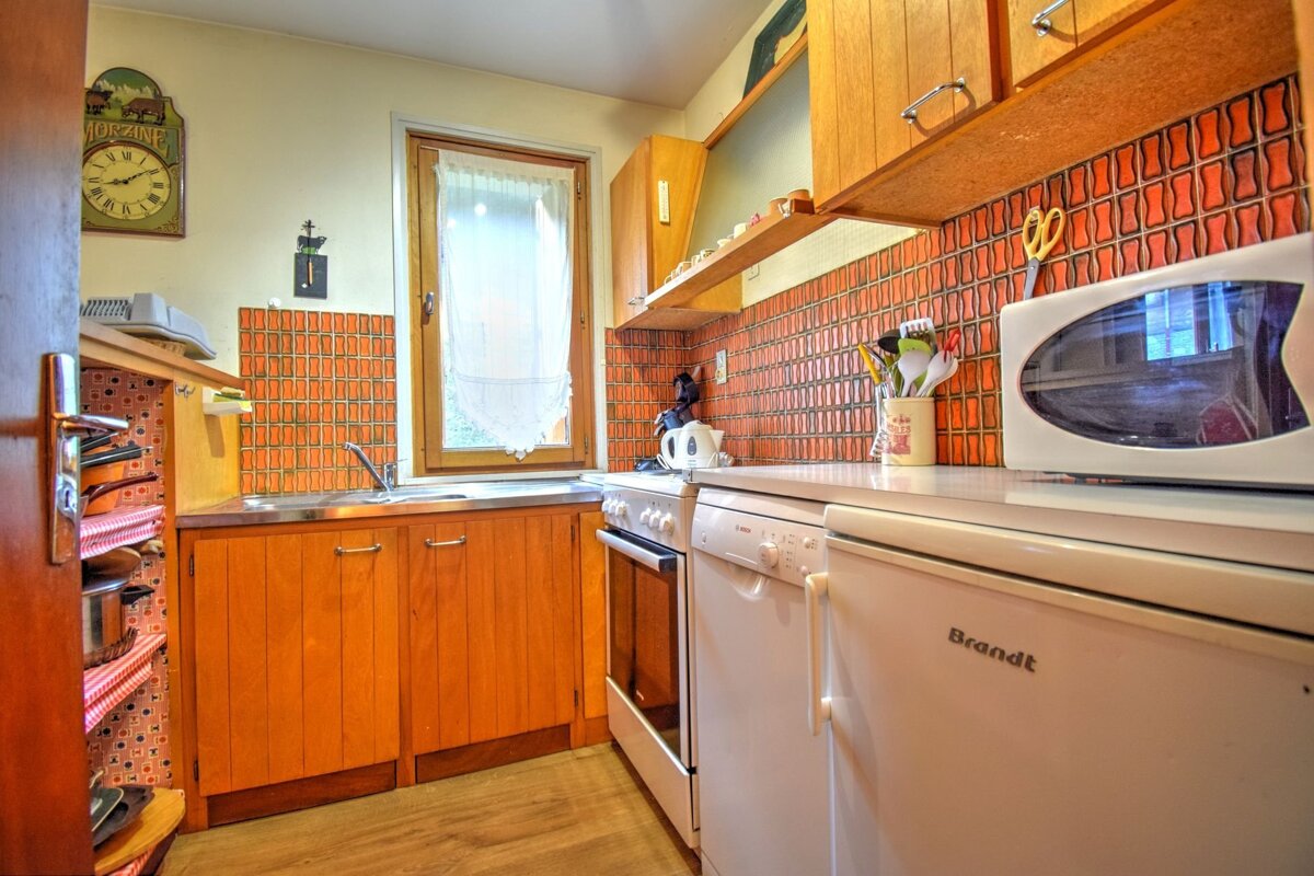 A retro kitchen with orange geometric tile backsplash, wooden cabinets, white appliances (fridge, oven, microwave), and a window.