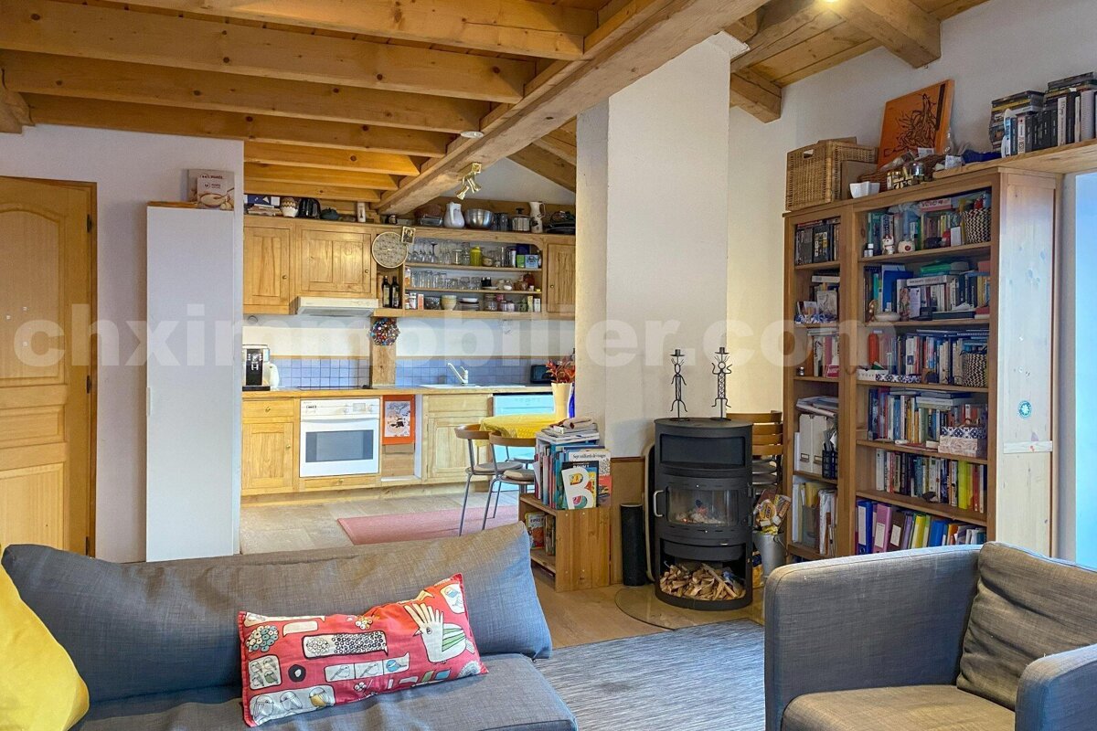 A cozy, open-plan living room and kitchen with wooden beams. Features a wood-burning stove, bookshelves, and rustic wooden kitchen cabinets.