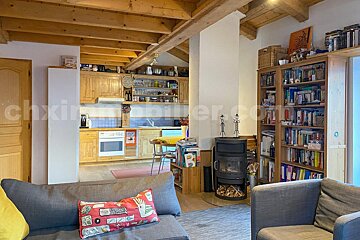 A cozy, open-plan living room and kitchen with wooden beams. Features a wood-burning stove, bookshelves, and rustic wooden kitchen cabinets.