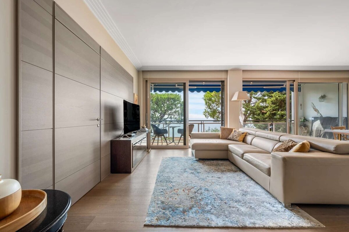 A modern living room features a beige sectional sofa, TV, wood wall, blue rug, and balcony with a view of trees and the sea.