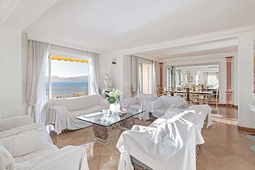 A sunlit, elegant living room with white slipcovered furniture, a glass table, and a balcony offering a sweeping sea view. A dining area is visible.