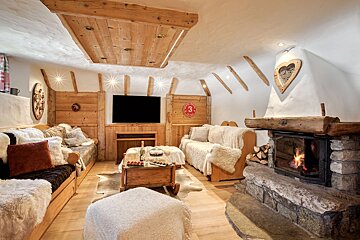 A warm, cozy chalet living room featuring a stone fireplace, wood-paneled walls and ceiling, plush sofas with fur throws, and a flat-screen TV.