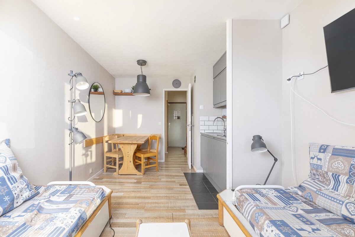 A bright studio apartment with two single beds, a wooden dining set, and a gray kitchenette. Light wood floors and modern decor fill the space.
