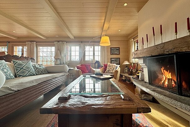 Cozy cabin living room with a roaring fireplace, large wooden coffee table, and comfortable seating under a wood-beamed ceiling.