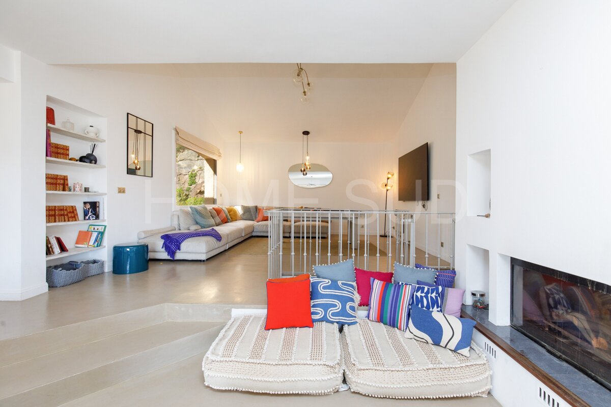 A bright, multi-level living room features a white L-shaped sofa with colorful cushions, built-in shelves, a large window, and floor seating by a fireplace.