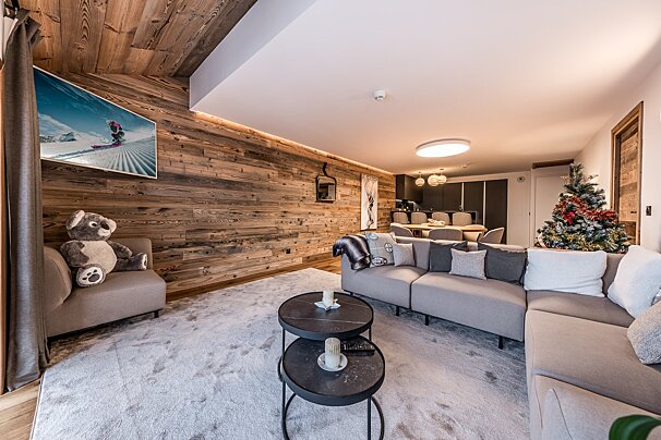 A modern, cozy living room with wood paneling, a large L-shaped sofa, a TV displaying skiing, and a Christmas tree, flowing into a dining area.