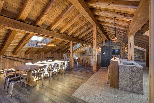 Rustic chalet interior with vaulted wood-beam ceilings, a long dining table with metal chairs, and a modern stone kitchen island.
