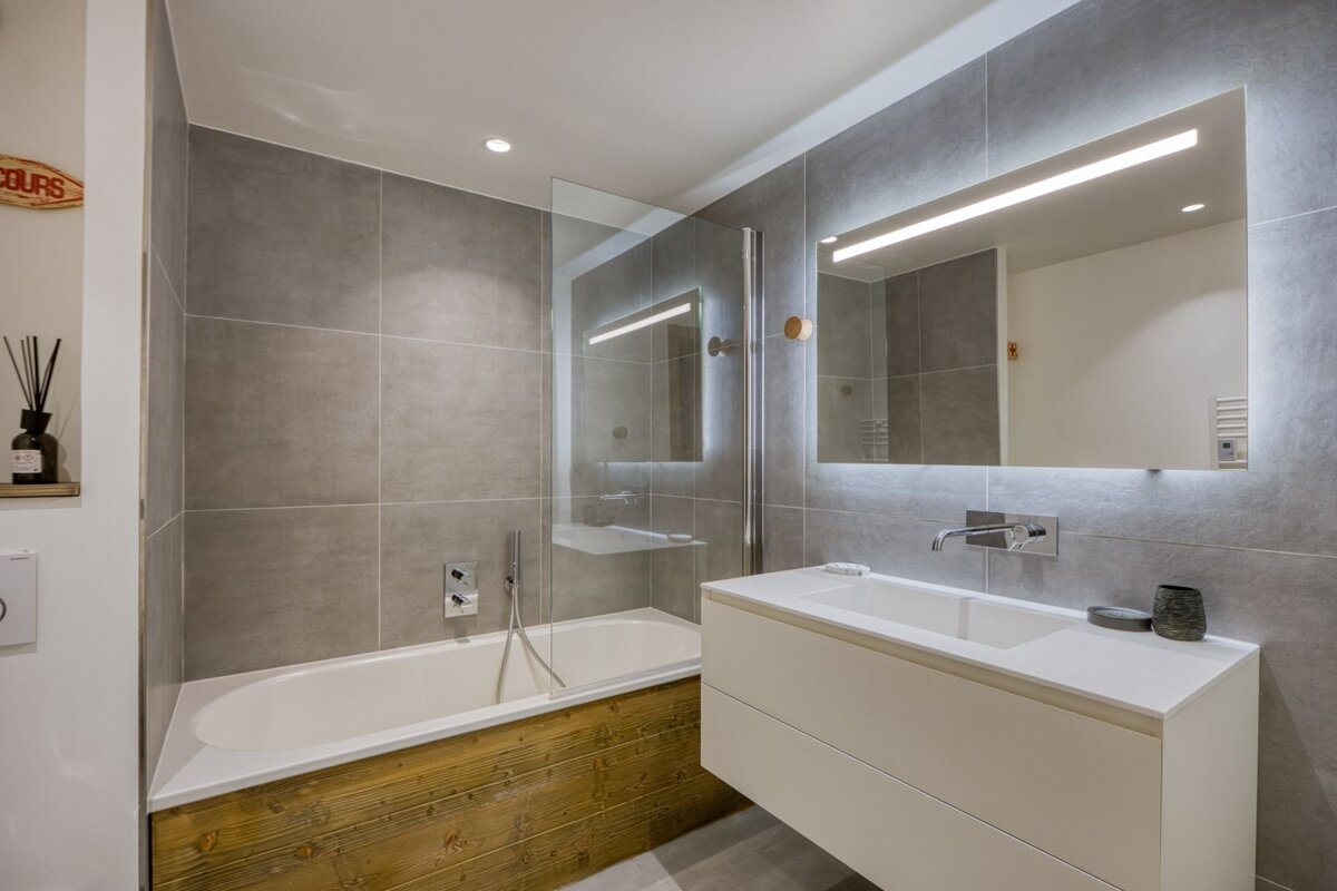 Modern grey-tiled bathroom featuring a white vanity with a backlit mirror, and a bathtub with a rustic wood-paneled side.