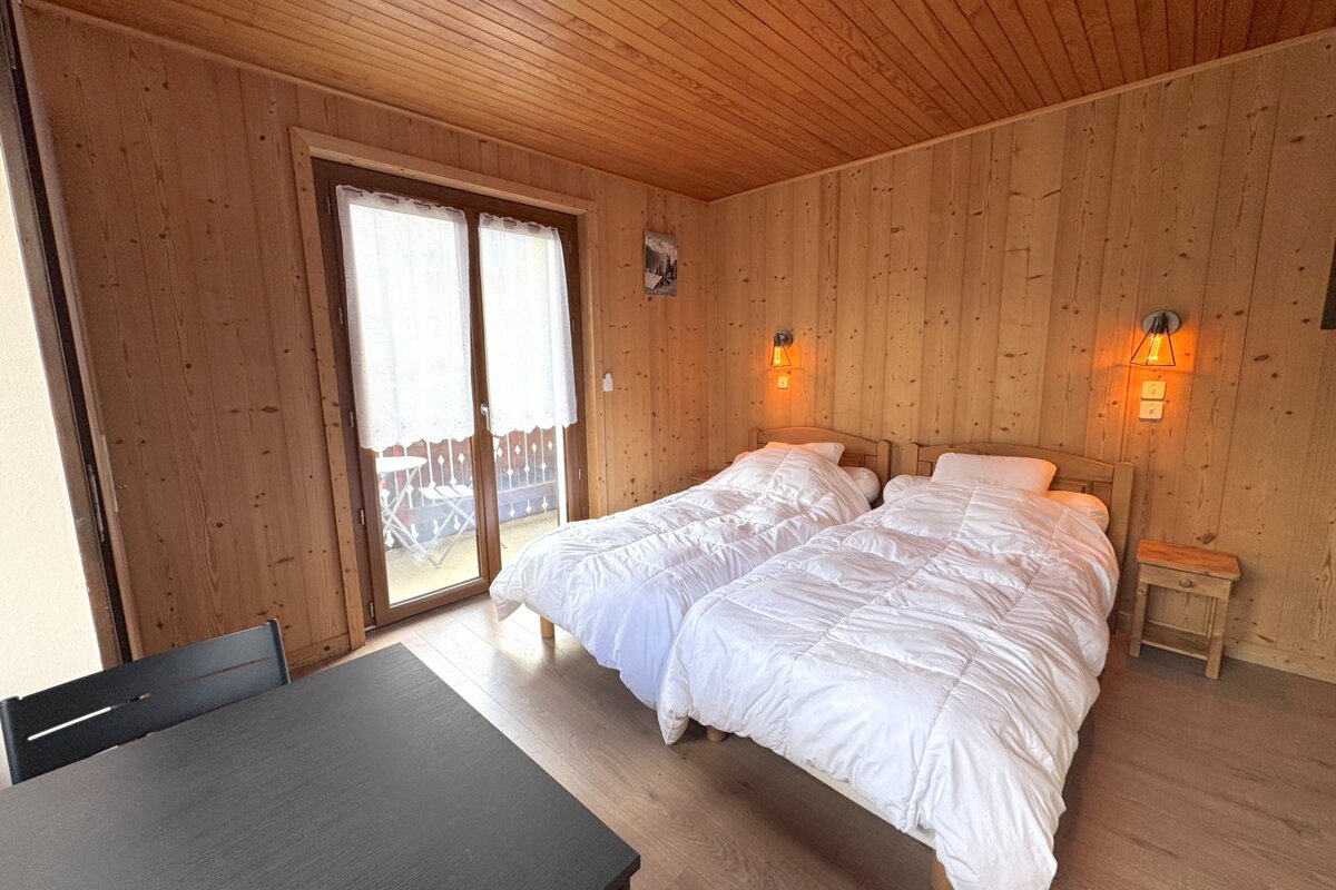 A cozy wood-paneled bedroom features two single beds with white duvets, warm wall lamps, and a glass door leading to a balcony.