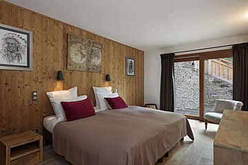 A rustic-modern bedroom featuring a wood-paneled wall, a large bed with red accent pillows, and artwork. A glass door opens to a stone outdoor area.